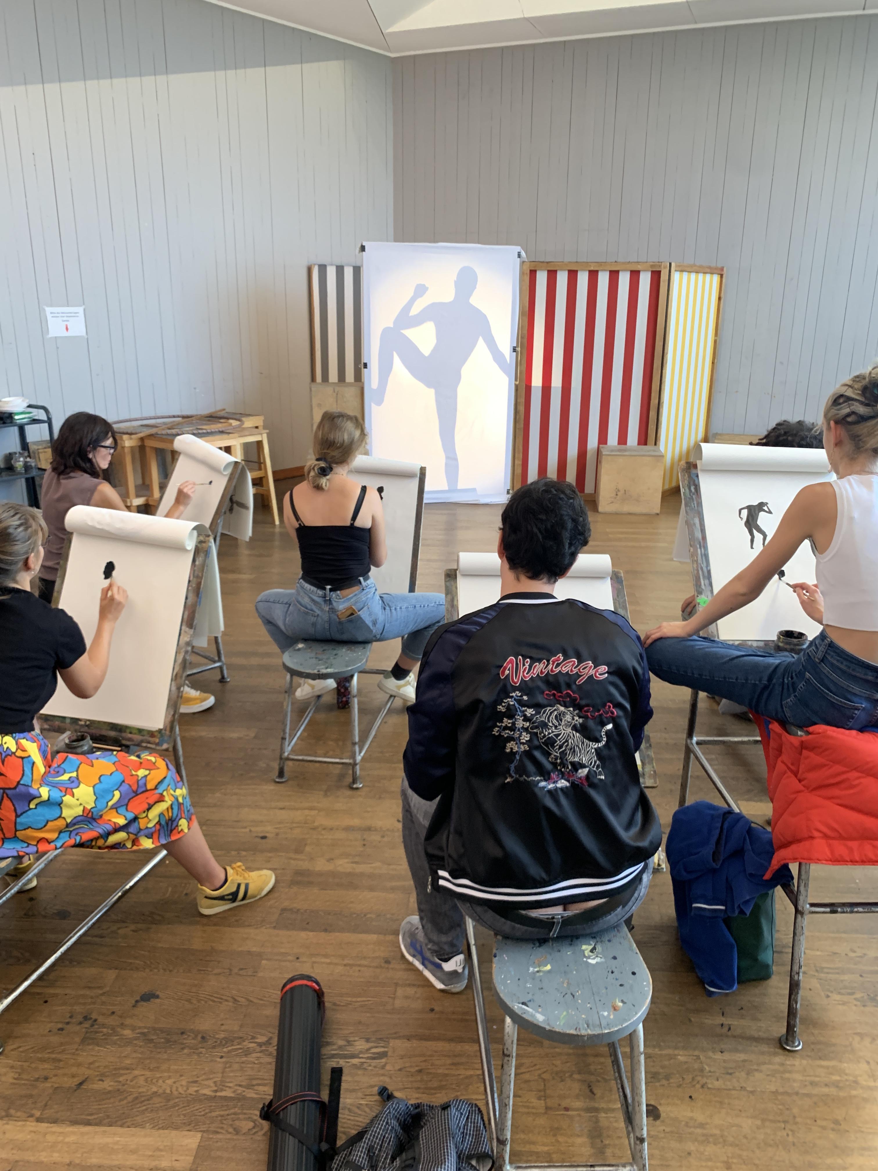 Art students seated in chairs with easels, sketching on canvases in a bright studio classroom