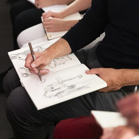A hand holding a pen drawing on paper against a black background